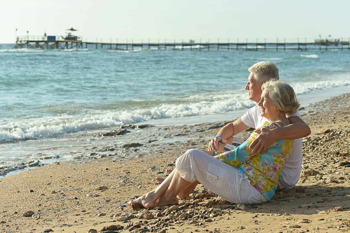 An older couple sits on the beach, perhaps thinking about end of life plans.